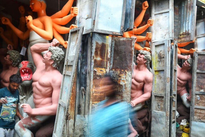 Large painted statues with multiple arms and muscular torsos are displayed behind old metal doors; a person in a blue shirt walks by, blurred in motion, in a busy, colorful workshop or market setting.