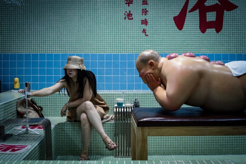 A woman in a tan dress and hat sits on a tiled bench, reaching into her bag. Next to her, a shirtless man lies face down on a massage table with cupping marks on his back in a tiled spa room with Chinese characters on the wall.