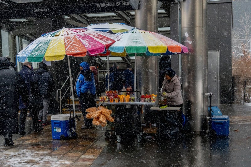 A street vendor stands under a colorful umbrella selling food and drinks during snowfall near a subway entrance; people in winter clothing walk by in the background.