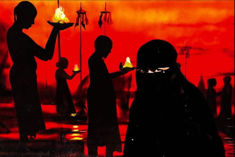 Silhouetted figures hold flaming bowls against a vivid red-orange background, with one person in the foreground wearing dark clothing and only their eyes visible. The scene has a dramatic, ceremonial atmosphere.