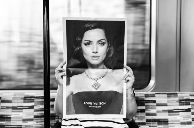 A person sitting on a patterned subway seat holds up a black-and-white Louis Vuitton jewelry ad, aligning the model's face in the ad with their own, creating the illusion of having the model's face.