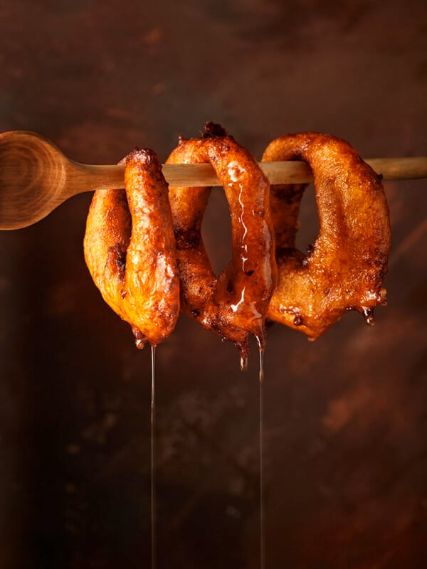 Three golden, deep-fried dough rings hang on a wooden spoon, glistening with syrup that drips down. The background is warm and softly blurred, emphasizing the texture and shine of the pastries.