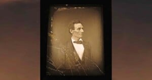 A cracked, sepia-toned photograph of a man in a suit with a bow tie, gazing to the side. The image appears old and weathered, with visible lines from broken glass across the surface.