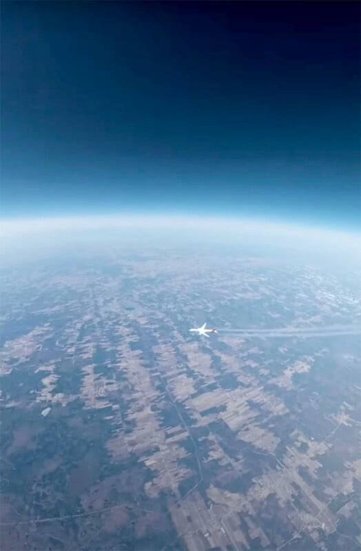 A high-altitude view of Earth shows a patchwork of fields and farmlands below a curved horizon, with a jet airplane flying across the landscape, leaving a visible contrail behind it under a deep blue sky.