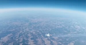 A commercial airplane flies high above a patchwork landscape, leaving a visible contrail behind, with the Earth’s curvature and blue sky in the background.