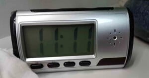 A digital clock displays the time 11:11. The clock has a silver frame with buttons labeled "HR," "MIN," "SET," "LIGHT" below the screen. It is placed on a white surface with a partially visible dark object nearby.