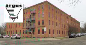 A large, red-brick, four-story industrial building on a street corner. It features multiple windows and a metal fire escape. Several parked cars line the street. An "Argus" logo with a geometric design is overlaid in the top left corner.