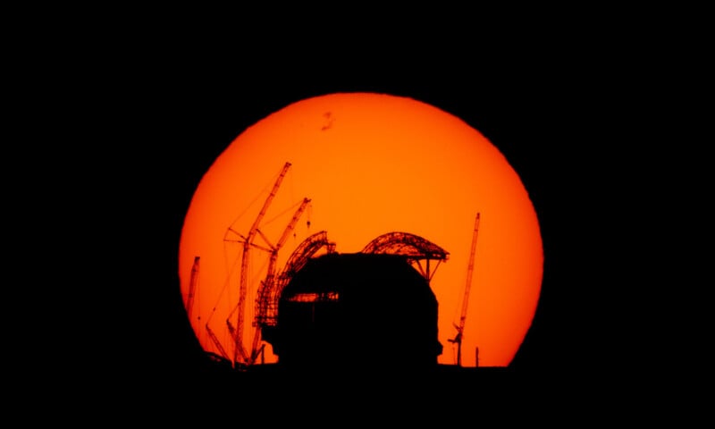 A large, bright orange sun dominates the background, silhouetting a building under construction with cranes and scaffolding, creating a striking contrast against the dark foreground.