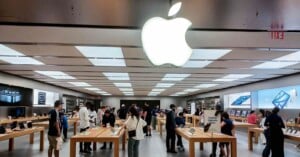 Inside an Apple Store, people browse products displayed on wooden tables. Large Apple logo is illuminated on the ceiling. Shelves filled with boxes and screens line the walls, and a busy atmosphere prevails with shoppers and staff interacting.