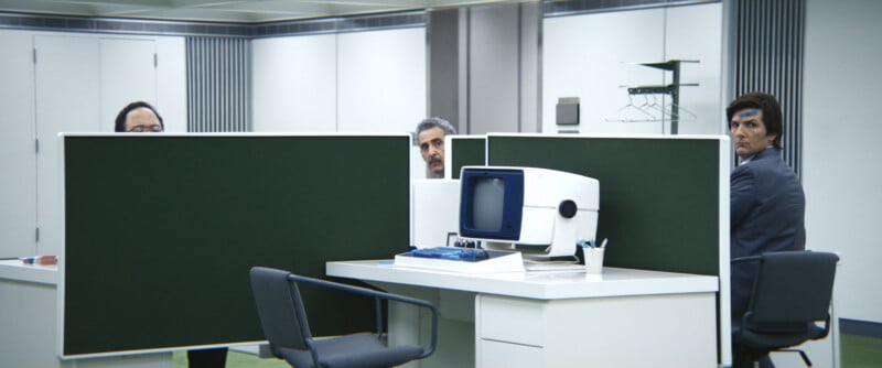 Three men sit at retro-style office desks separated by high green dividers, looking up from their work. A vintage computer and office supplies are on the desks. The setting has a minimalist, mid-century modern feel.