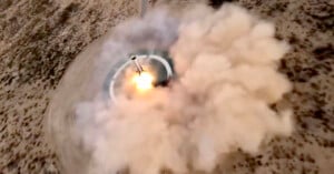 A rocket launches from a circular pad in a desert area, producing bright flames and a large cloud of smoke that spreads over the surrounding sandy landscape.