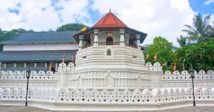 A white-walled temple with a red-tiled roof and small flags, surrounded by decorative walls and greenery, under a partly cloudy sky.
