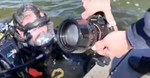 A scuba diver wearing a full-face mask is partially submerged in water. Nearby, a person is holding a Sony camera close to the diver's face, possibly taking a photo or video. The water is slightly rippled and appears greenish.