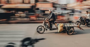 A man rides a motorized tricycle with a cart attached on a city street, carrying bags. The background and another biker are motion-blurred, emphasizing speed and movement.