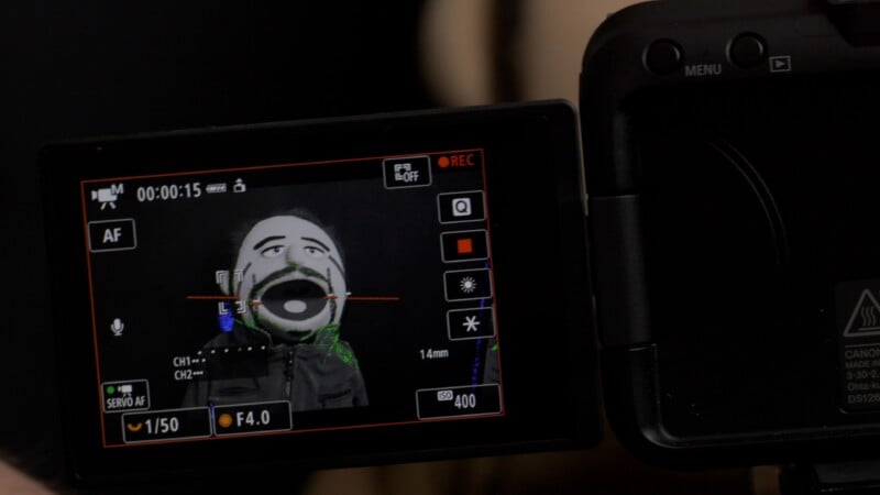 Close-up of a camera screen displaying a puppet with dark hair and an open mouth. The screen shows various camera settings, such as focus, aperture, and ISO. The camera body is partially visible to the right.