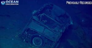 A rusted, partially buried vehicle rests on the ocean floor, captured in deep blue water with "Ocean Exploration" and "Previously Recorded" labels on the image.