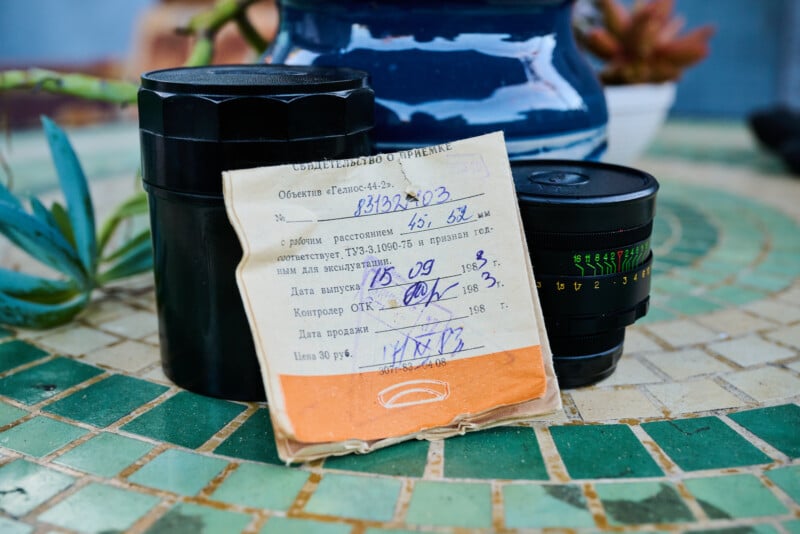 A camera lens with its black case and a vintage, handwritten instruction sheet rests on a green-tiled mosaic table. A blue vase and some plants are blurred in the background.