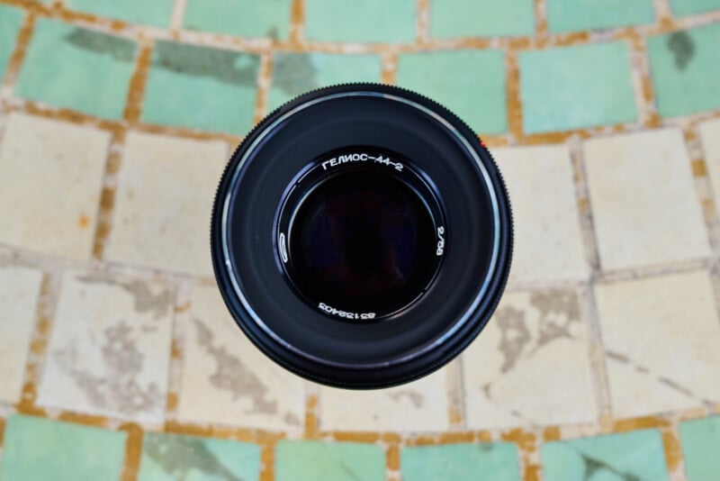 A black camera lens with Cyrillic writing is photographed from above, centered against a tiled background of alternating green and beige squares.