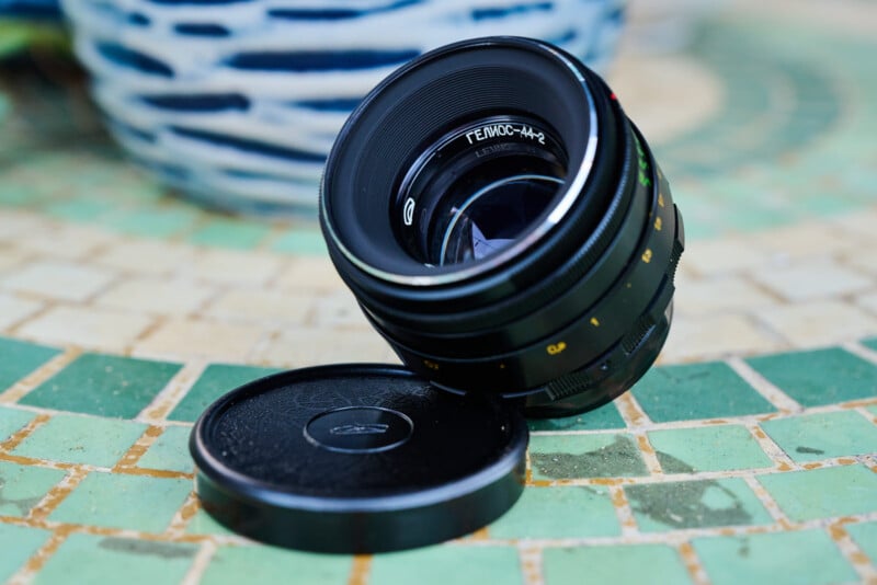 A camera lens with Cyrillic writing on it is standing at an angle on a mosaic tiled surface, next to its detached lens cap. The background is blurred.