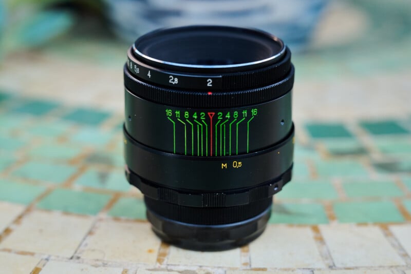 A close-up of a black camera lens with green, white, and red markings on its barrel, placed on a patterned surface with a blurred background.