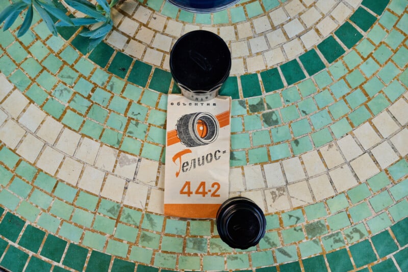 A vintage camera film canister and a box labeled “ФЭД” (FED) with “44-2” lie on a circular, green-and-white mosaic tile surface. A plant is partially visible in the corner.