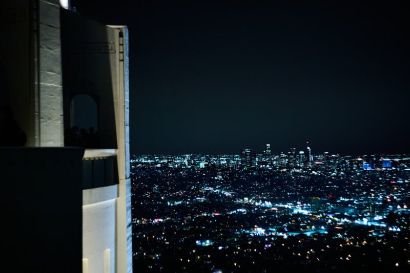 A nighttime cityscape photo showing the brightly lit skyline of Los Angeles, with the illuminated white tower of the Griffith Observatory on the left side of the image.