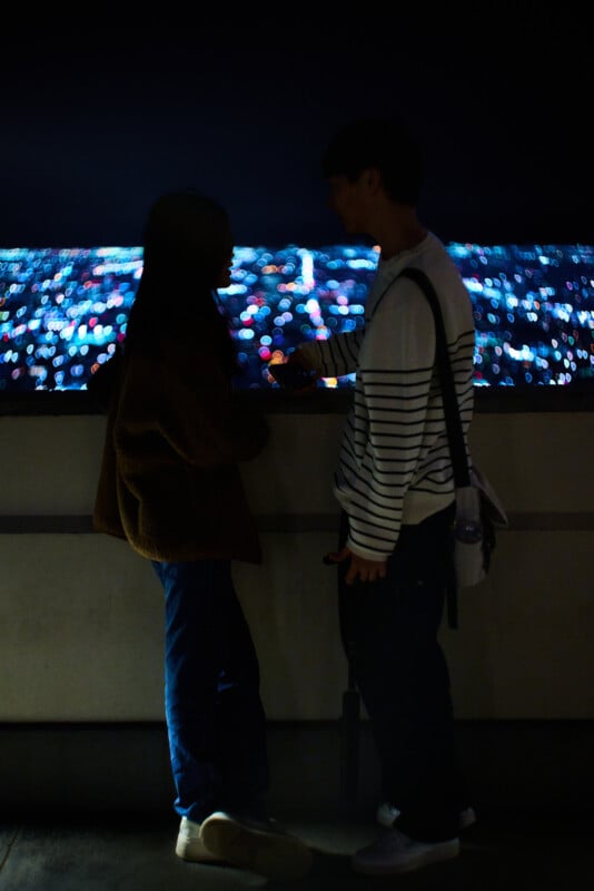 Two people stand close together at night, silhouetted against a cityscape filled with blurred, colorful lights in the distance. They appear to be talking or sharing a quiet moment.