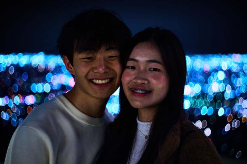 A young couple smiles together at night, with colorful, out-of-focus lights creating a bokeh effect in the background.