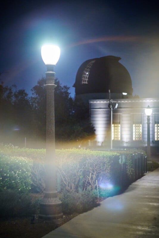 A glowing streetlamp illuminates a nighttime scene with a distant domed observatory building in the background, surrounded by bushes and trees. A bright light flare and soft glow add a dreamy effect to the image.