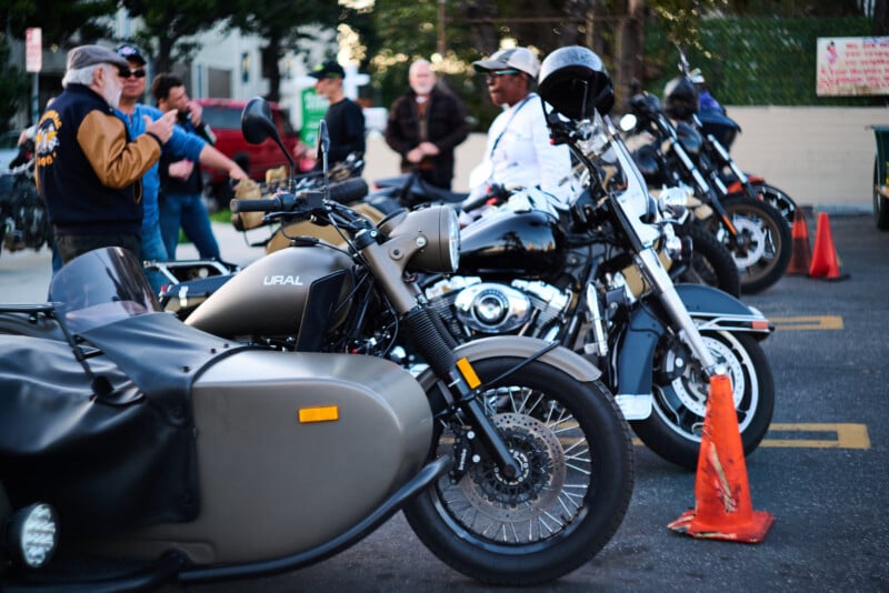 Several motorcycles, including one with a sidecar, are parked in a lot. Orange traffic cones are nearby, and a group of people stands in the background, talking and wearing casual or motorcycle gear.