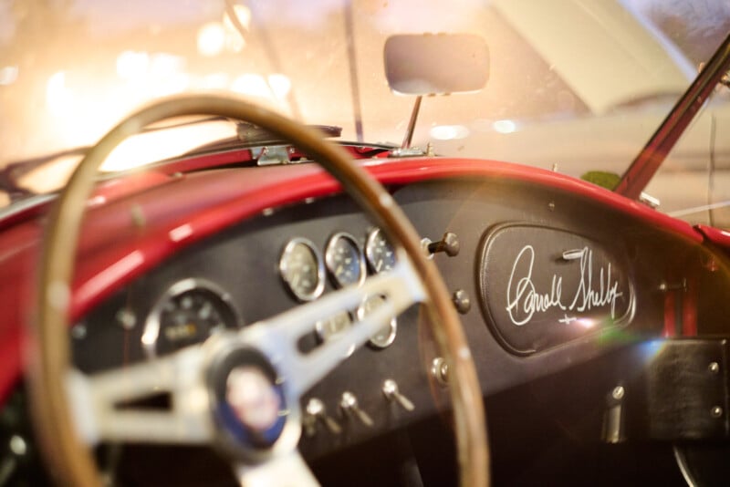 Close-up view of the interior of a classic red car, showing a wooden steering wheel, vintage dashboard gauges, and a signature that reads "Carroll Shelby" on the glove compartment.