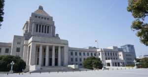 A large stone government building with a central pyramid-shaped tower, columns at the entrance, and a Japanese flag on a flagpole, surrounded by trees under a clear blue sky.