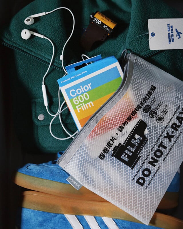 A close-up of travel essentials: a green fleece, white earphones, a roll of 35mm film, a Polaroid Color 600 film pack, blue Adidas sneakers, and a mesh pouch labeled "FILM DO NOT X-RAY" with text in English and Chinese.