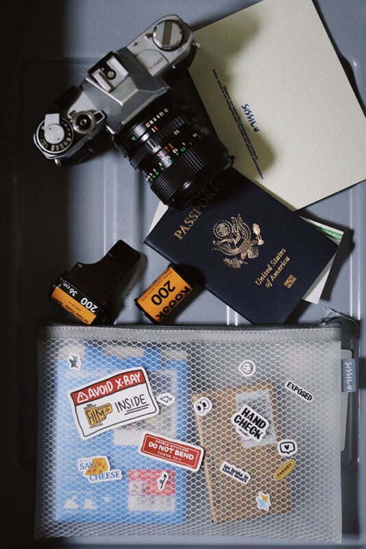 A film camera, a roll of 200 ISO film, a U.S. passport, a document, and a mesh bag with various stickers are placed inside a gray security bin, likely for airport screening.