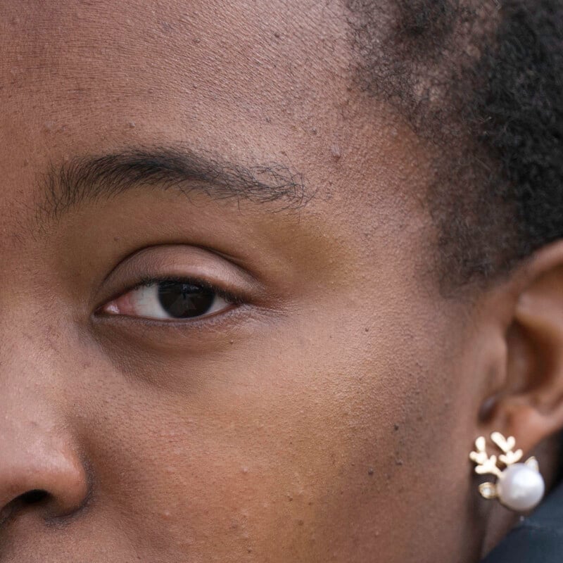 Close-up of the left side of a person's face, focusing on their eye, eyebrow, forehead, and ear. The person is wearing a gold earring with a pearl and a branch-like design. Their skin texture and hairline are visible.