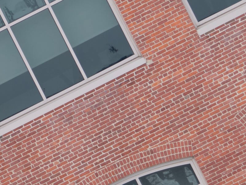 A tilted image of a red brick building facade featuring large rectangular windows with white frames. The bricks are uniformly arranged, and the windows reflect a cloudy sky. The building has a modern appearance with clean lines.