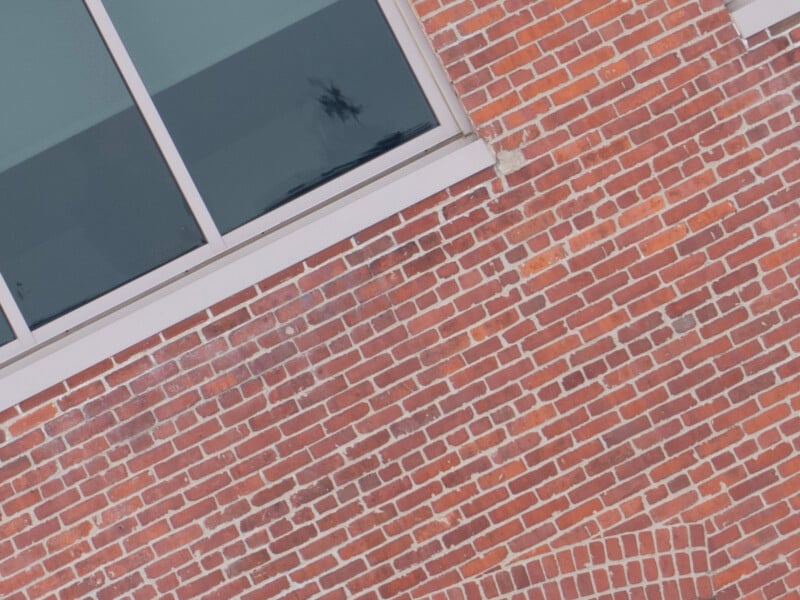 A section of a brick wall with a window in the top left corner. The bricks are red with white mortar, and part of a wooden window frame is visible. The photo is slightly tilted, giving a dynamic angle to the composition.
