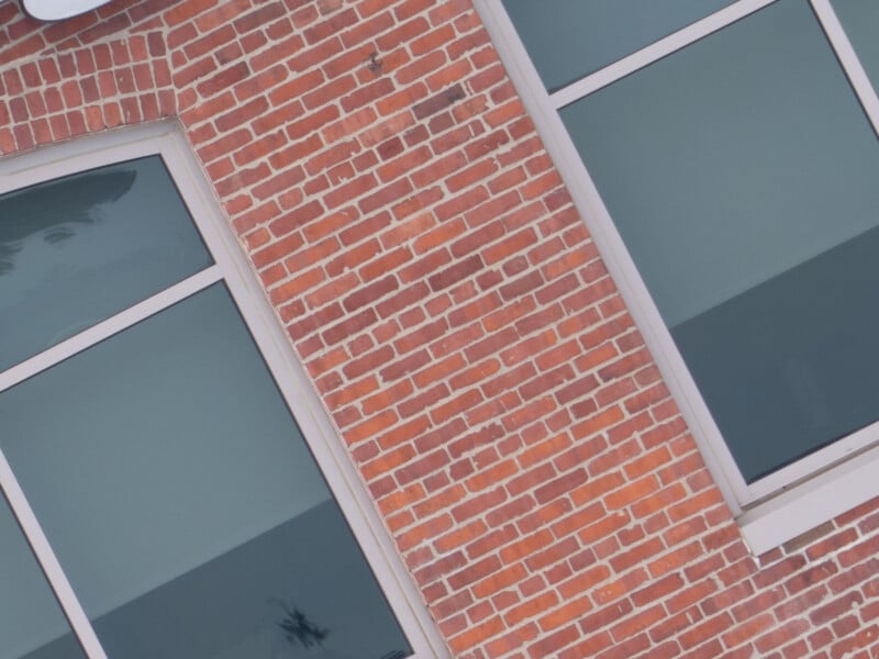 A diagonal view of a red brick wall with three large rectangular windows. The windows have blue-tinted glass, reflecting the light. The upper left corner shows a small portion of a white overhang or structure.