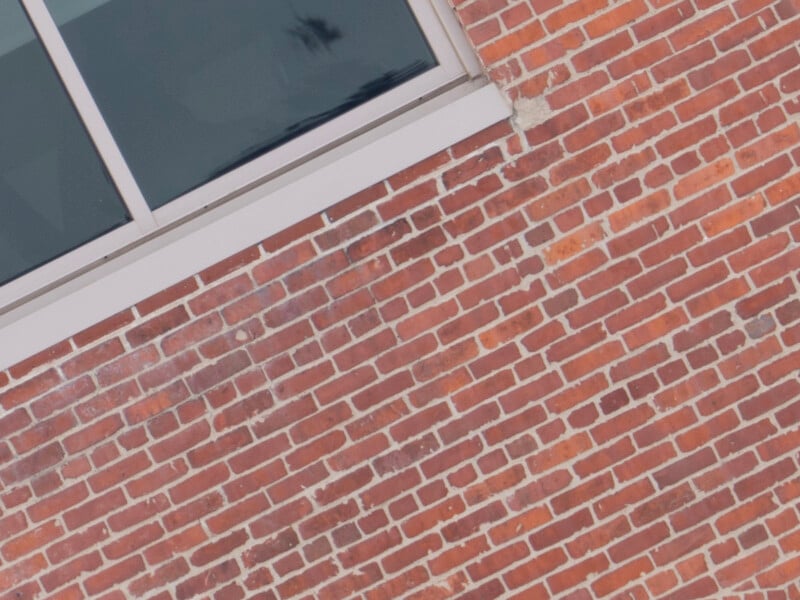 A section of a red brick wall with a white-framed rectangular window positioned near the top left corner. The image is tilted, showing the textured bricks and a partial glimpse of the cloudy sky reflected in the window.
