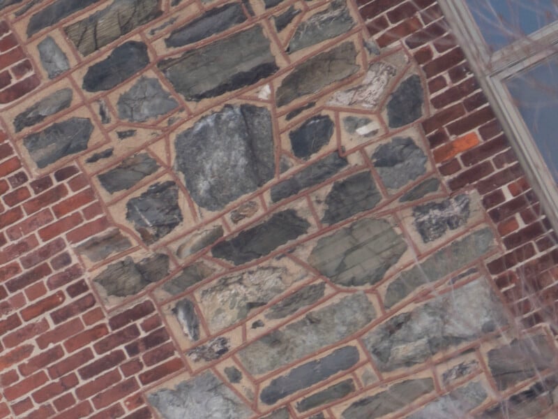 Close-up of a brick and stone wall, featuring irregularly shaped gray stones set into a brick facade. The stones vary in size and are surrounded by mortar. A portion of a window is visible on the right side of the image.