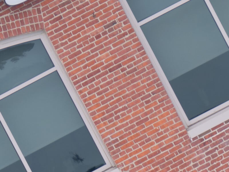 A close-up of a brick wall with two rectangular windows. The bricks are red-orange with white mortar, and the windows have white frames. Both windows reflect some exterior elements, creating a slight glint.