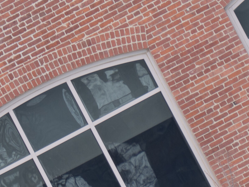 Close-up of a red brick building facade with large, arched windows. The window panes reflect an abstract pattern of nearby structures, and the brickwork features a curved arch above the window frame.