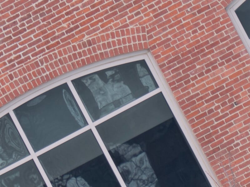 A close-up of a brick building with two large windows. The windows have a white frame and are reflecting a cloudy sky and tree branches. The brick pattern is consistent, and the building's corner is slightly visible.
