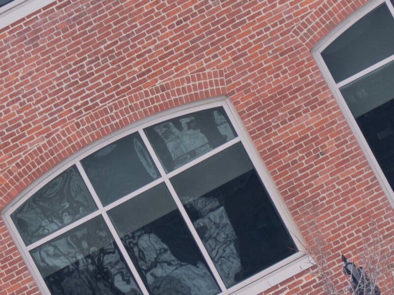 A close-up view of a red brick wall featuring large, arched windows with white frames. The windows reflect a distorted image of the outside landscape, while some leafless branches are slightly visible at the bottom right corner.