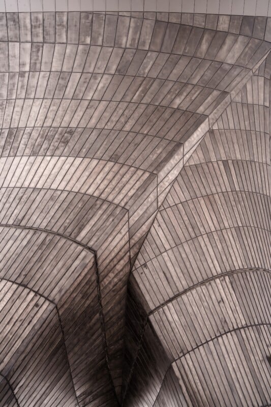 Abstract architectural detail featuring intersecting, curved surfaces covered with a grid pattern of rectangular metal tiles. The play of light and shadows enhances the geometric complexity, creating a visually striking composition.