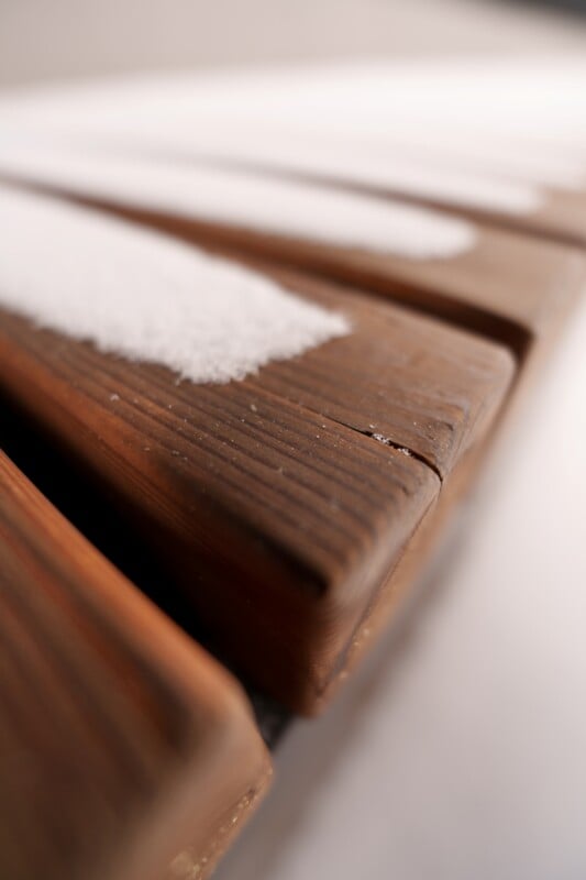 Close-up of a wooden surface with strips of snow resting on it, showing distinct lines between the snow and the exposed wood. The focus is on the texture and detail of the wood grain and the contrast with the snow.