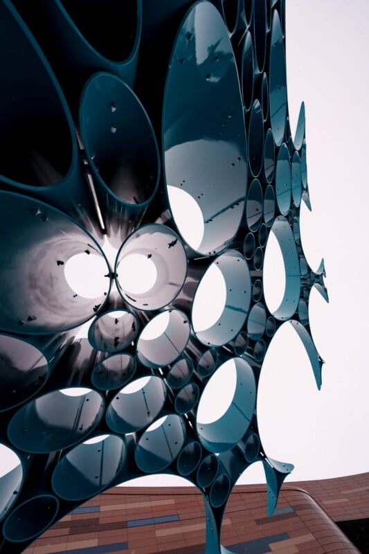 A close-up view of a modern sculpture composed of intersecting metal tubes with circular openings, creating an intricate pattern. The tubes are dark and reflective, and the background shows a building with a geometric facade.