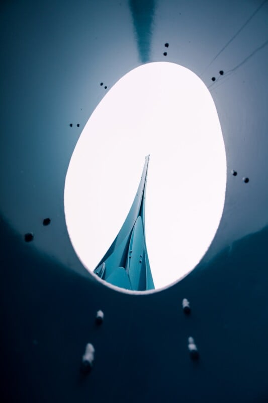 View of a sail through an oval opening in a dark surface. The abstract composition highlights the contrast between the smooth, blue surface of the sail and the surrounding darker border.