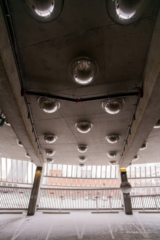 Underground parking garage with a concrete ceiling featuring round lights. Lines of lights create a pattern. The garage has multiple empty parking spaces, and a snowy scene is visible outside through the metal railing.