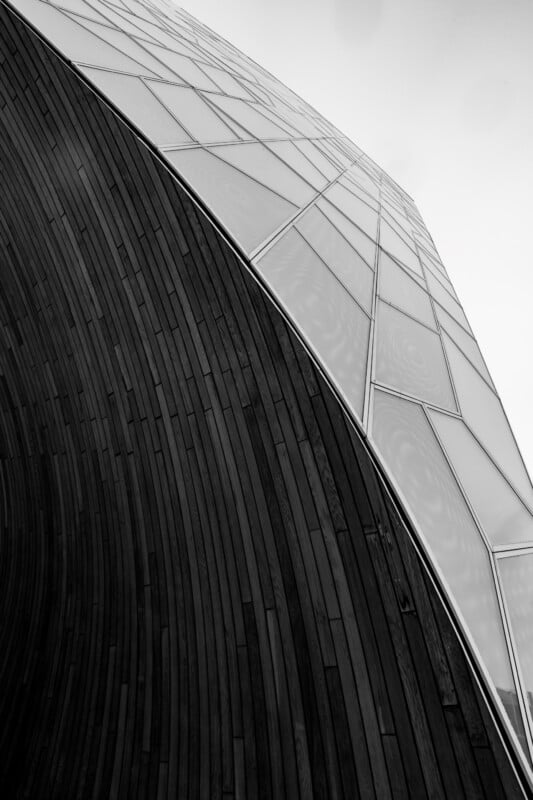 Close-up of a modern architectural structure with a curved wooden wall on the left and a geometric glass facade on the right, blending textures and materials under a grey sky.